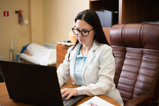Female doctor wearing a lab coat and glasses, focused on working with a laptop at her desk, with a patient resting on an examination bed in the background of the clinic room