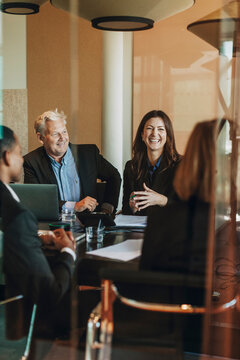 Happy female law expert sitting with colleagues in meeting room at office seen from glass