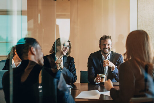Smiling male and female lawyers sitting with colleagues in meeting room at office seen from glass