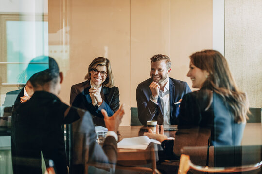 Smiling male and female business professionals sitting with colleagues in meeting room at law office seen from glass