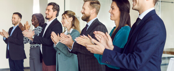 Diverse happy smiling young business people in suits standing in a row on conference and listening to colleague or leadership. Company employees applauding on meeting in office. Banner.