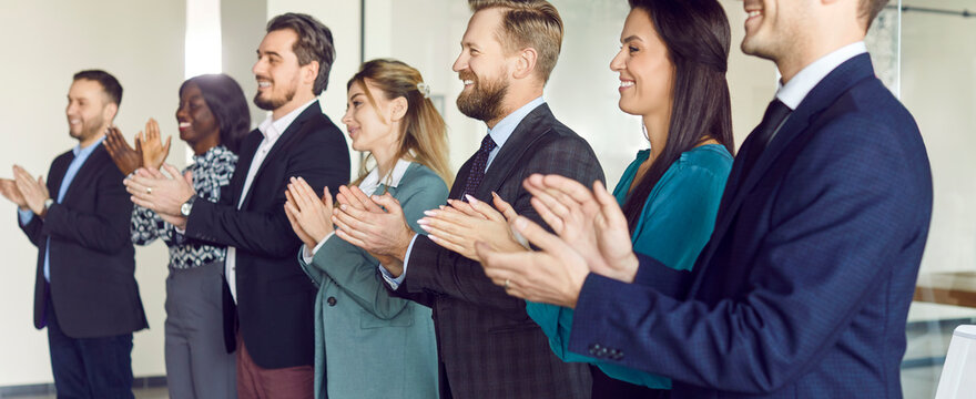 Diverse happy smiling young business people in suits standing in a row on conference and listening to colleague or leadership. Company employees applauding on meeting in office. Banner.