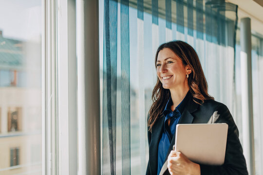 Smiling female law professional holding laptop looking away while standing in office corridor