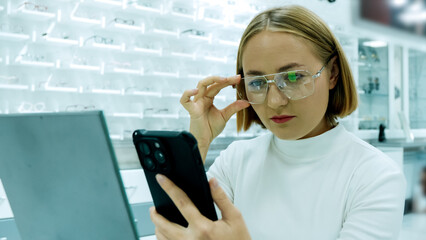 A woman is wearing glasses and looking at her smartphone in an optical store