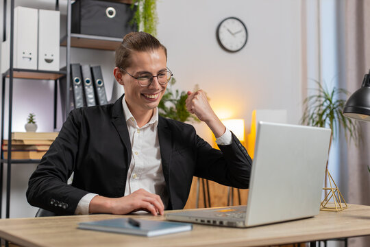 Happy young Caucasian businessman working on laptop netbook shocked by sudden good career news, victory, winning, goal achievement. Excited freelancer guy clenching fists at home office workplace.