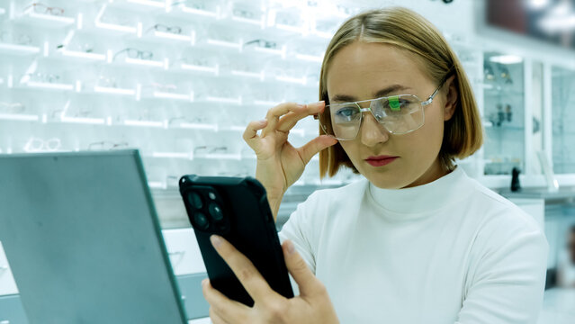 A woman is wearing glasses and looking at her smartphone in an optical store - Powered by Adobe
