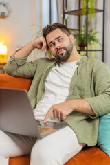 Overtired Caucasian young man with brown hair bored using laptop while sitting on sofa at home....