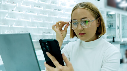 woman is trying on a pair of glasses while looking at her smartphone in a shop
