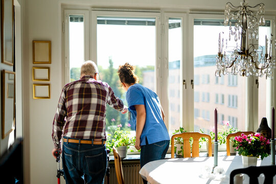 Rear view of elderly man and female nurse looking out window in living room at home