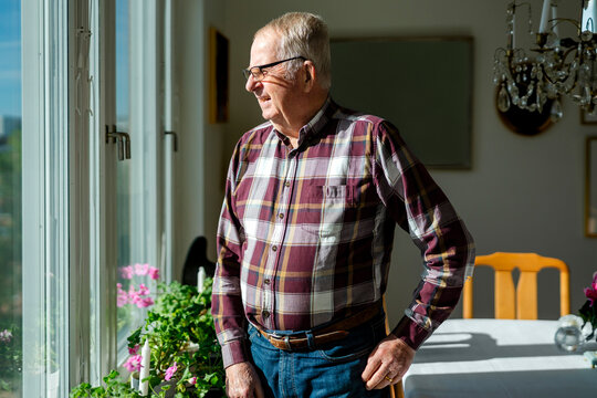 Senior man looking out window while standing in living room at home