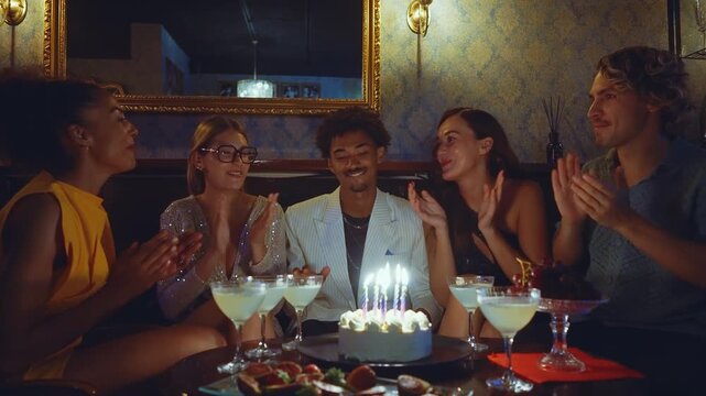 Joyful multi-ethnic group of friends celebrating a birthday party in a fancy bar, clapping and cheering while a happy young man makes a wish and blows out the candles on the cake
