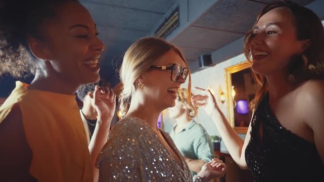 Happy diverse women in sparkling outfits dancing and socializing at a nightclub. Group of girlfriends enjoying a fun night out, celebrating together in a lively bar with music and dim lights