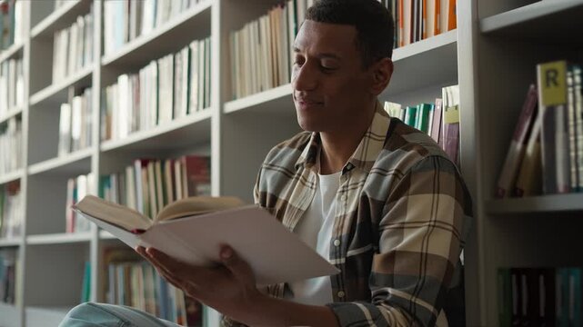 Calm African American man student reading book sitting on library flor bookshelves ethnic male guy enjoying literature learning read story knowledge university studying bookworm focused reader hobby