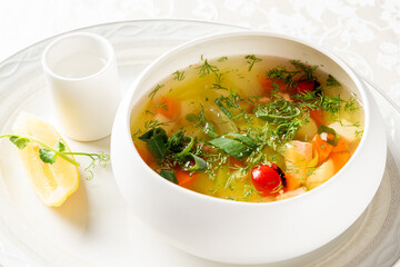 A bowl of healthy, clear vegetable or chicken soup with fresh herbs, carrots, and tomatoes on a white table setting with lemon wedge