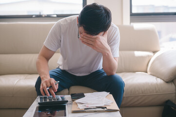 Stressed man reviewing bills and expenses at home while managing financial problems. A worried man sitting on a couch holding bills, stress rising expenses, financial difficulties, debt management