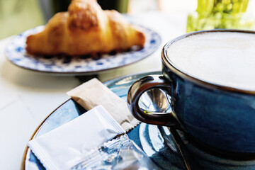 Latte in blue cup with sugar packets and croissant on marble table in cozy café setting
