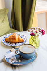 Latte with heart foam and croissant on marble table with flowers in cozy café setting