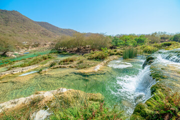 Wadi Darbat natural park in Dhofar, Oman, presenting clear emerald water with waterfalls cascading over lush rocks and green trees under a bright blue sky