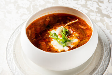 A hearty, vibrant red borscht soup in a white bowl, garnished with a swirl of sour cream and fresh green microgreens on a light patterned tablecloth