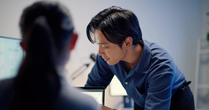 A close-up, side-view portrait of a serious Asian businessman leaning over a desk, holding a pen and discussing data on a computer monitor with a colleague in a dim, late-night office.