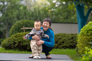 An Asian grandmother in a blue top crouches on a narrow path, holding her 16-month-old grandson in plaid shirt close, with green foliage in the background