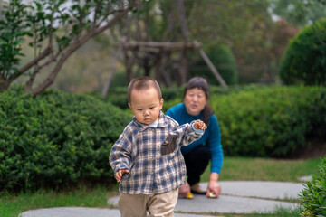 An Asian man and grandmother watching a 16-month-old baby boy run in a park, the grandmother in a blue top and baby in plaid shirt