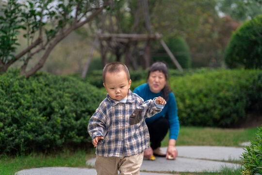 An Asian man and grandmother watching a 16-month-old baby boy run in a park, the grandmother in a blue top and baby in plaid shirt - Powered by Adobe
