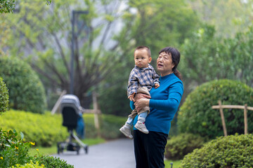 An Asian grandmother in a blue top, holding her 16-month-old grandson against a green botanical background - minimalist and tender moment