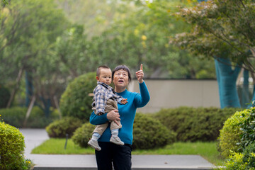 An Asian grandmother in a blue top, holding her 16-month-old grandson against a green botanical background - minimalist and tender moment