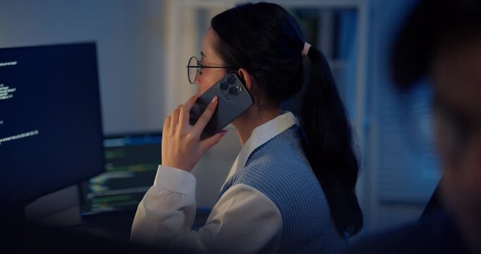 A close-up side profile of a professional Asian businesswoman wearing glasses, making a business phone call while continuing to work on her computer screen late alone in a dark office.