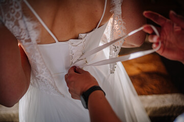 Bride in elegant white gown is having her dress laced up by a helper, showcasing intricate details...