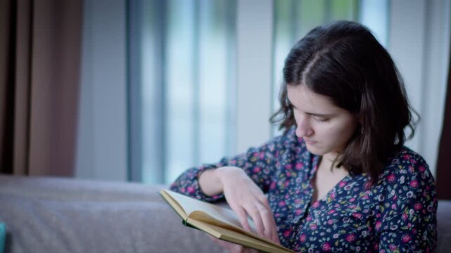 A young woman spends her time reading a book while sitting on the sofa at home