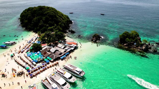 An aerial drone view of Koh Khai Nok in Thailand, showing turquoise water, crowded sandy beaches, colorful umbrellas, and speedboats anchored along the shore