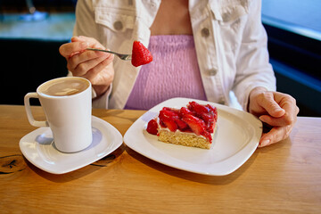 A woman enjoys a slice of strawberry cake with a cup of coffee, lifting a fresh strawberry on her fork at a cozy café table.