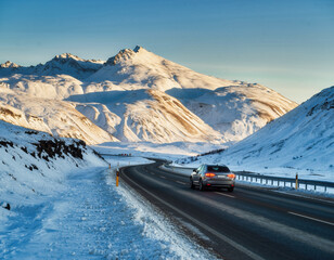 Car drives along snowy mountain road under clear blue sky, surrounded by majestic snow covered peaks
