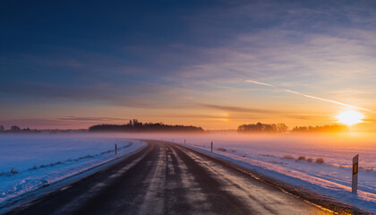 Serene winter sunrise casts warm glow over frosty rural road, with misty fields and clear sky