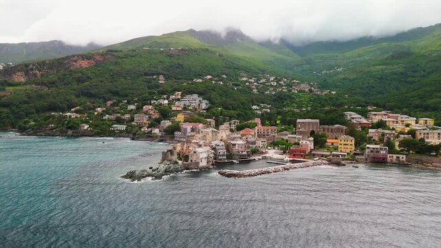 Smooth Aerial Approach Shot Flying Over Mediterranean Sea Towards Erbalunga Fishing Village, Highlighting the Historic Genoese Tower Ruins and Colorful Port Houses, Cap Corse, Corsica, France.