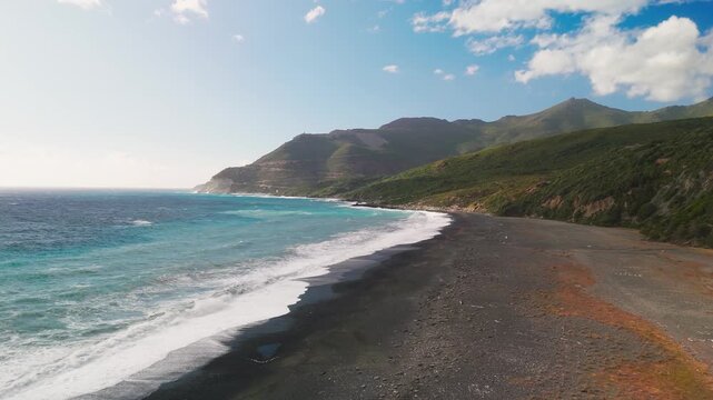 Low altitude aerial drone flight over the unique dark grey sand of deserted Plage de Nonza beach, contrasting with the lush green Cap Corse landscape and blue Mediterranean Sea, Corsica, France.