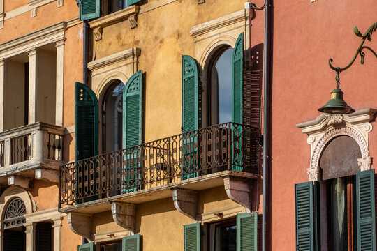 Close-up of a colorful historic building facade in Verona, Italy, with a decorative iron balcony, arched windows and green wooden shutters opened to the warm afternoon sun. The textured yellow and ter - Powered by Adobe