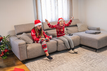 Sisters wearing santa hats and festive sweaters enjoying playing video games on a sofa in their living room on christmas day