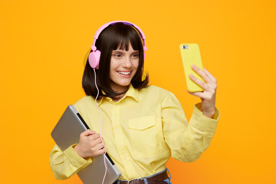 Cheerful student wears a pastel yellow shirt and pink headphones while posing for a selfie, holding a notebook and laptop; a bright backdrop conveys modern lifestyle, creativity, and youthful energy.