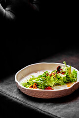 A close-up, dimly lit photo of a fresh gourmet green salad with avocado, nuts, and dressing served on a white textured bowl, set against a dark background.