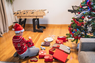 Young boy in a festive sweater and santa hat opening gifts on the floor next to a decorated christmas tree