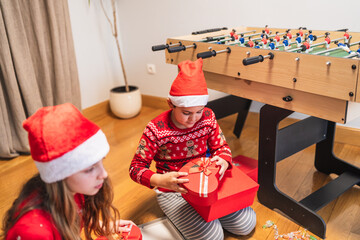Young siblings wearing santa hats and festive sweaters eagerly unwrapping gifts by a foosball...