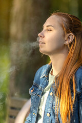 An young woman with long, red hair vaping in a forested area. The person is captured from the side, exhaling a cloud of vapor from an electronic cigarette. Summer landscape in a background.