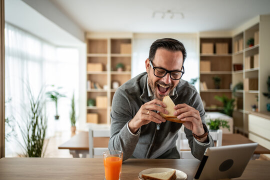 Cheerful man eating sandwich and watching digital tablet at home
