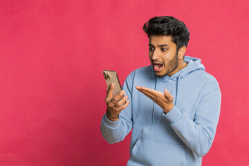 Upset sad Indian man use smartphone typing browsing, play game, loses, surprised by sudden lottery results bad fortune loss fail unlucky news. Arabian Hindu bearded guy isolated on red background