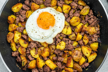 Beef hash with fried potatoes, ground beef and sunny egg in skillet on gray surface with dark napkin, perfect for rustic cooking themes.