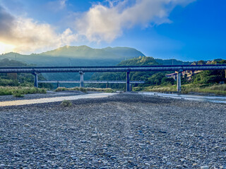 Wide view of a dry gravel riverbed beneath an elevated viaduct bridge in Wulai, New Taipei City, Taiwan. Mountains are illuminated by bright morning sunlight against a blue sky.