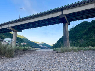 Looking up at the massive concrete structure of an elevated viaduct from the rocky riverbed. Green hills and village buildings visible in the distance on a clear morning. Wulai, Taiwan.